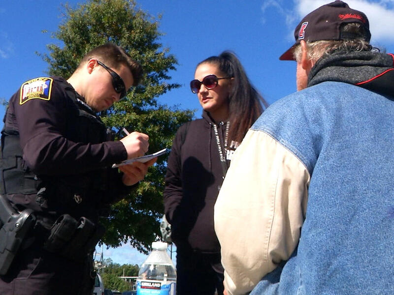A police officer writes in a notebook while speaking with a woman in sunglasses. A man with his back to the camera in a baseball hat is also part of the conversation.