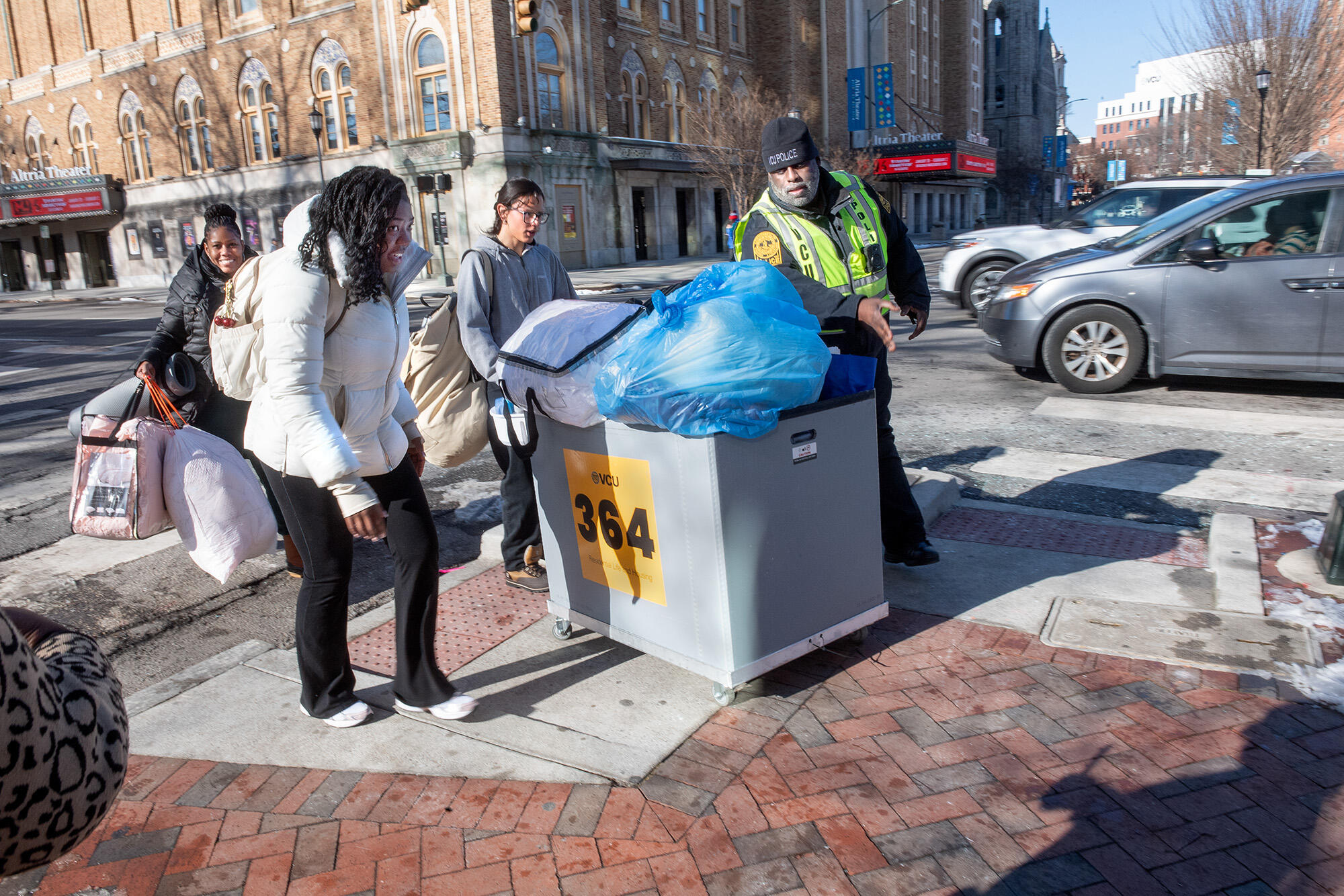 A photo of a VCU Police officer standing to the right of one woman who are carrying bags, one woman is is walking and wearing a back pack, and one who is pushing a large bin.