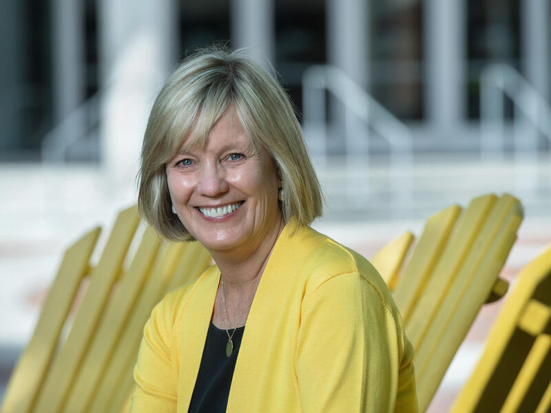 A photo of a woman from the chest up. She is smiling and sitting in front of a row of three yellow chairs. 
