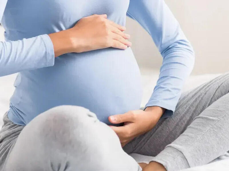 A photo of a woman from the neck down. She is sitting with her legs crossed and her hands cupping the top and bottom of her pregnant stomach. 