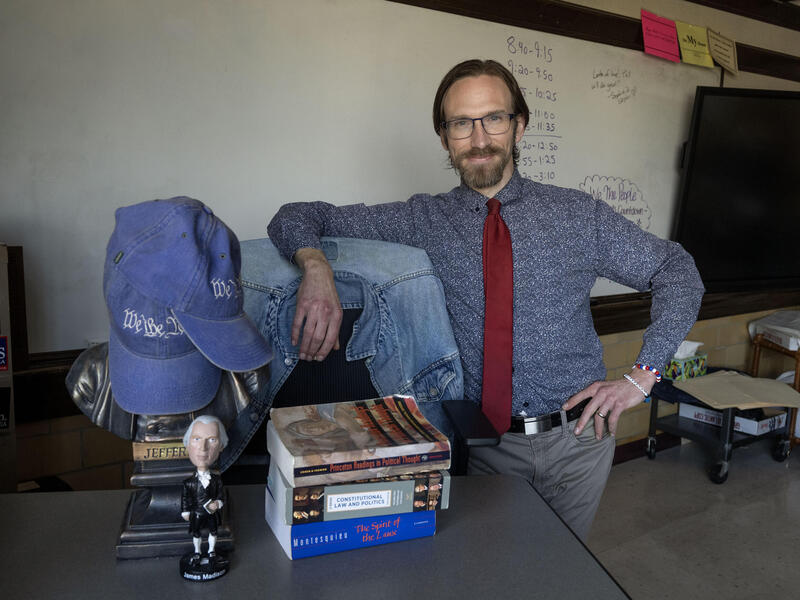A photo of a man leaning his left arm against a chair and his right hand on his hip. He is standing next to a desk that has a stack of books, a bobblehead of James Madison, and a bust of Thomas Jefferson with two denim baseball caps that say \"We the People\" in white text covering its face. 