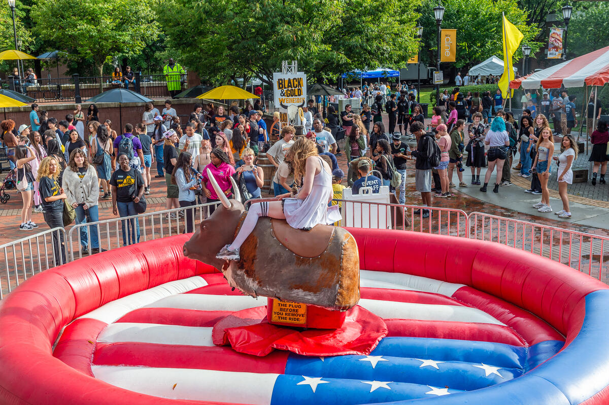 A photo of a person riding a mechanical bull that is set up in the middle of an inflatable ring. The bull set up is surrounded by a gate. Behind the gate are people walking around 