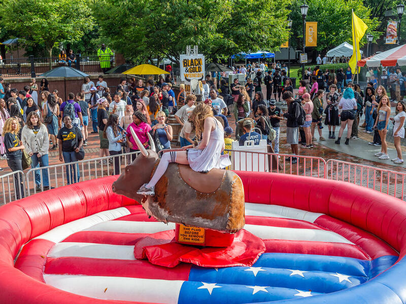 A photo of a person riding a mechanical bull that is set up in the middle of an inflatable ring. The bull set up is surrounded by a gate. Behind the gate are people walking around 