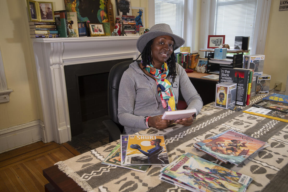 A woman sitting at a desk with comic books on it. She is holding a book with both of her hands. 