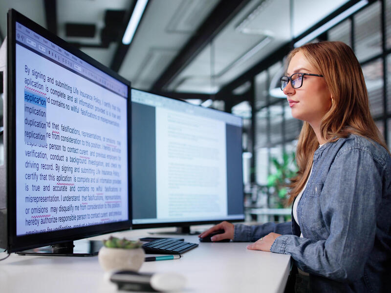 A photo of a woman sitting at a desk with two computer monitors on it. She is looking at the monitor on the left which is covered in text. 