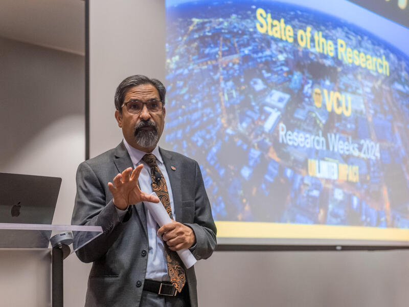 A man in a coat and tie speaks and gestures with his right hand while standing in front of a video screen.