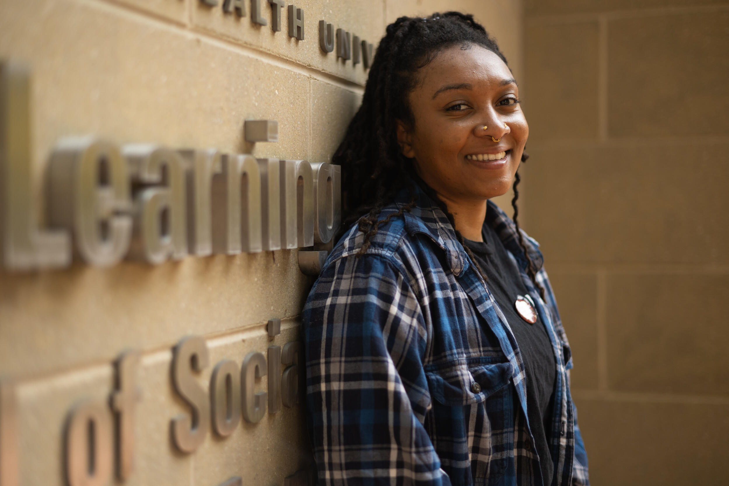A woman in a flannel shirt smiles and leans against a wall.
