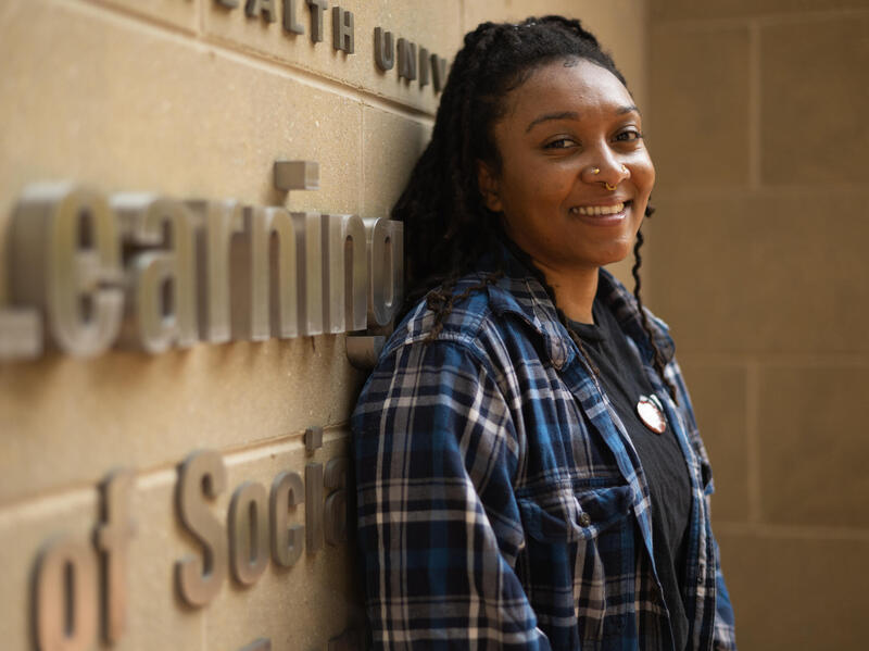 A woman in a flannel shirt smiles and leans against a wall.