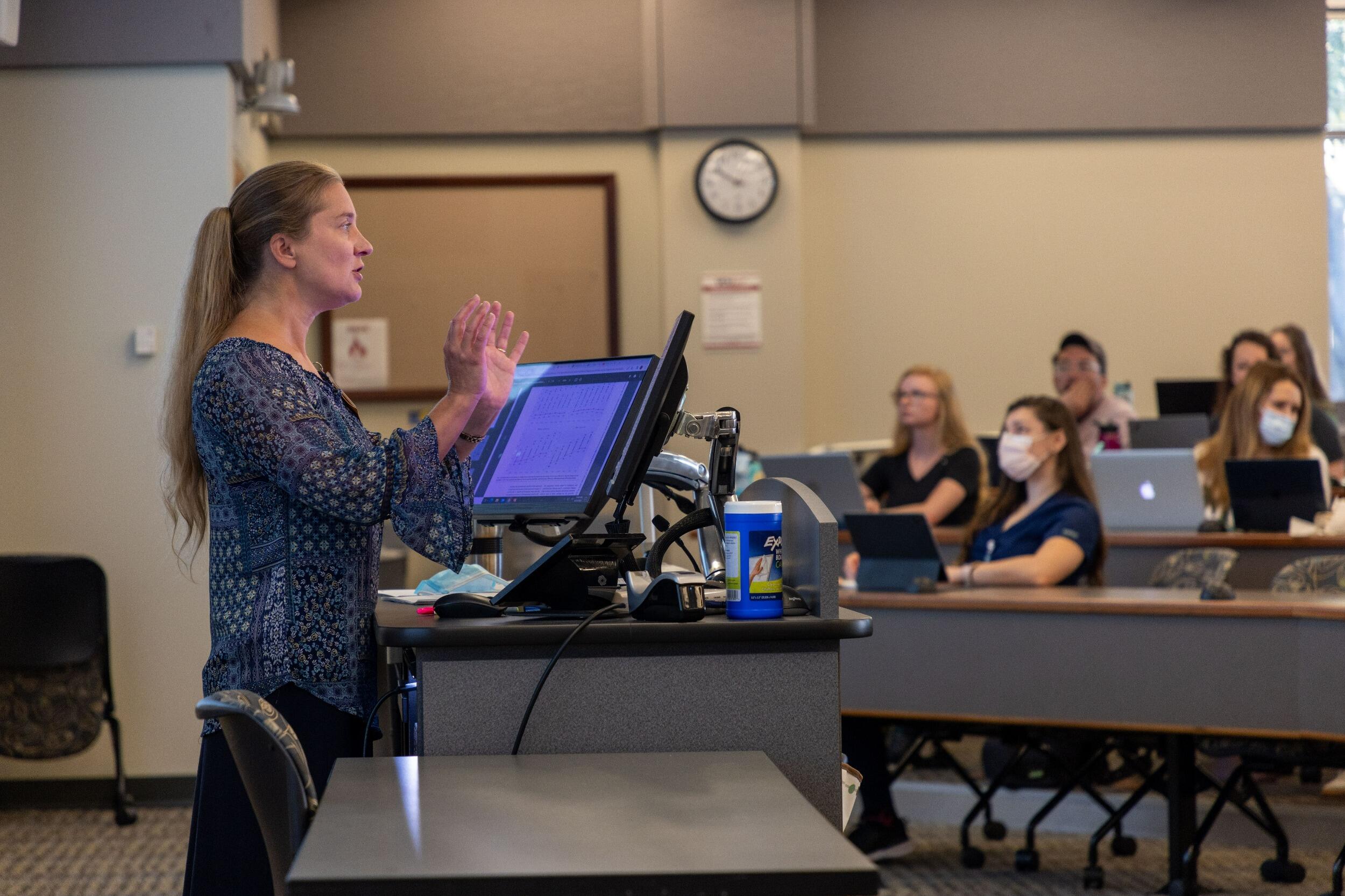 A woman speaking at a podium in front o fo a room full of students. 