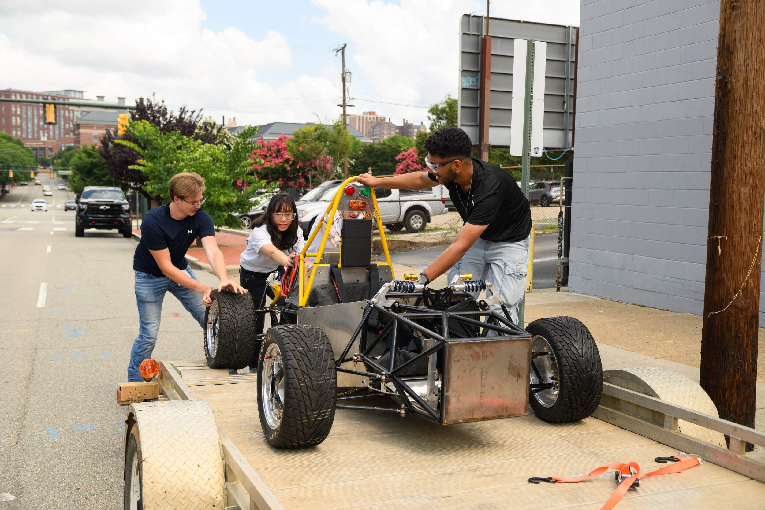 A photo of three people wheeling a car frame off of a trailer bed. 