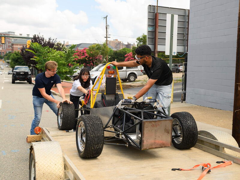 A photo of three people wheeling a car frame off of a trailer bed. 