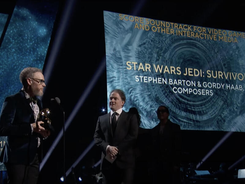 A photo of two men standin on a stage. The man on the left is standing holding an award while the man on the right watches him. Behind them is a screen that reads \"STAR WARS JEDI: SURVIVOR STEPHEN BARTON & GORDY HAAB COMPOSERS) 