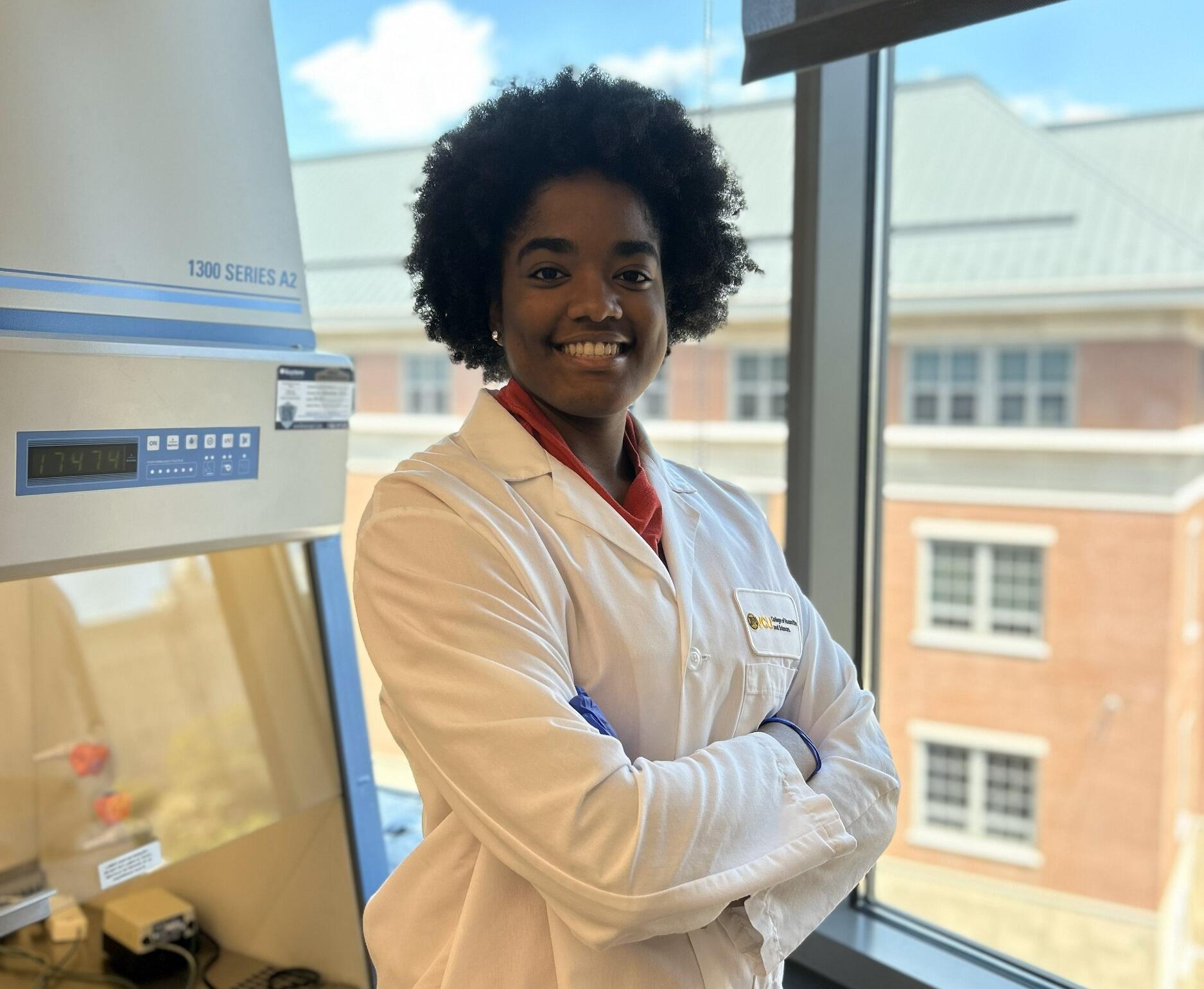 A woman standing in front of a window with her arms crossed against her chest. She is wearing a white lab coat and smiling. 