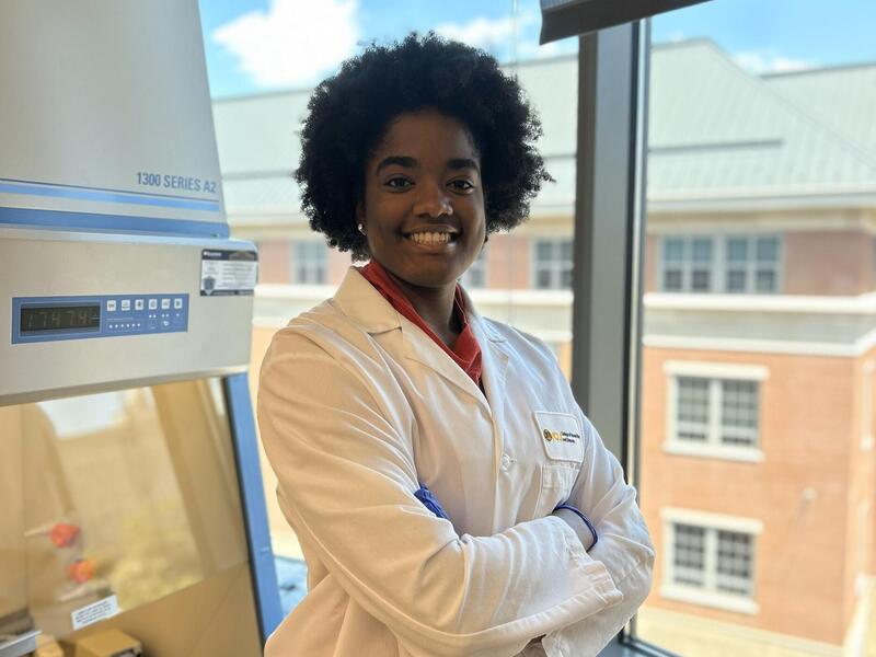 A woman standing in front of a window with her arms crossed against her chest. She is wearing a white lab coat and smiling. 