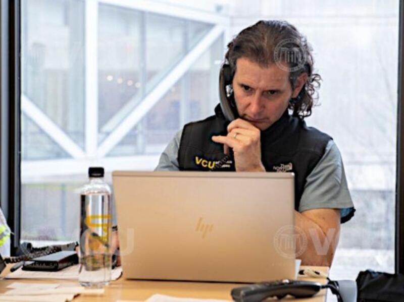 A man sitting in front of a laptop looking at the screen while holding a phone against his ear. 