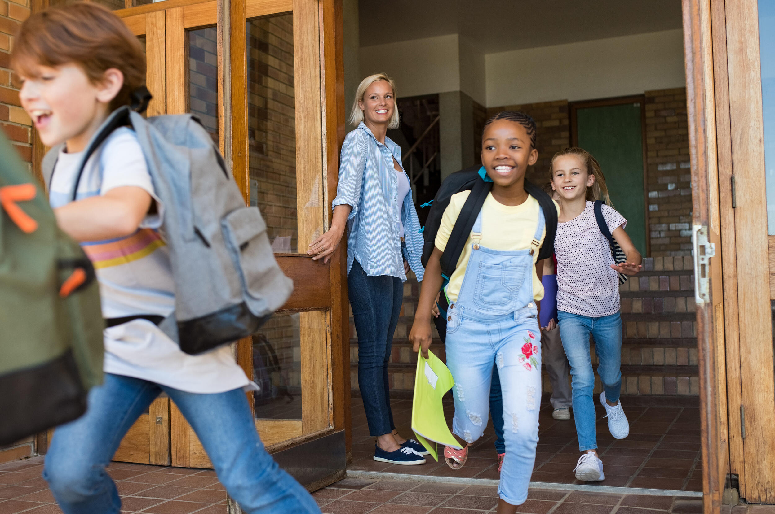 A photo of children running out of a school. A teacher is holding the door for them and smiling. 
