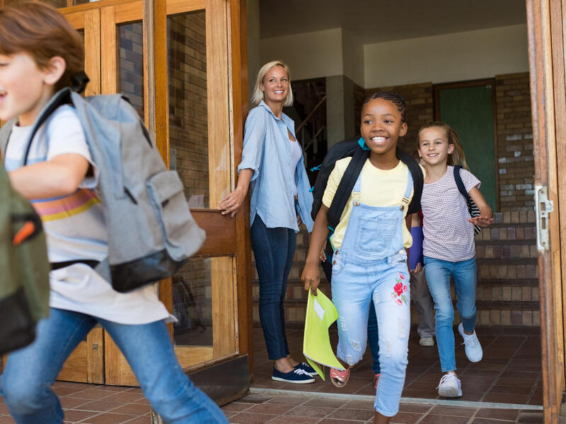 A photo of children running out of a school. A teacher is holding the door for them and smiling. 