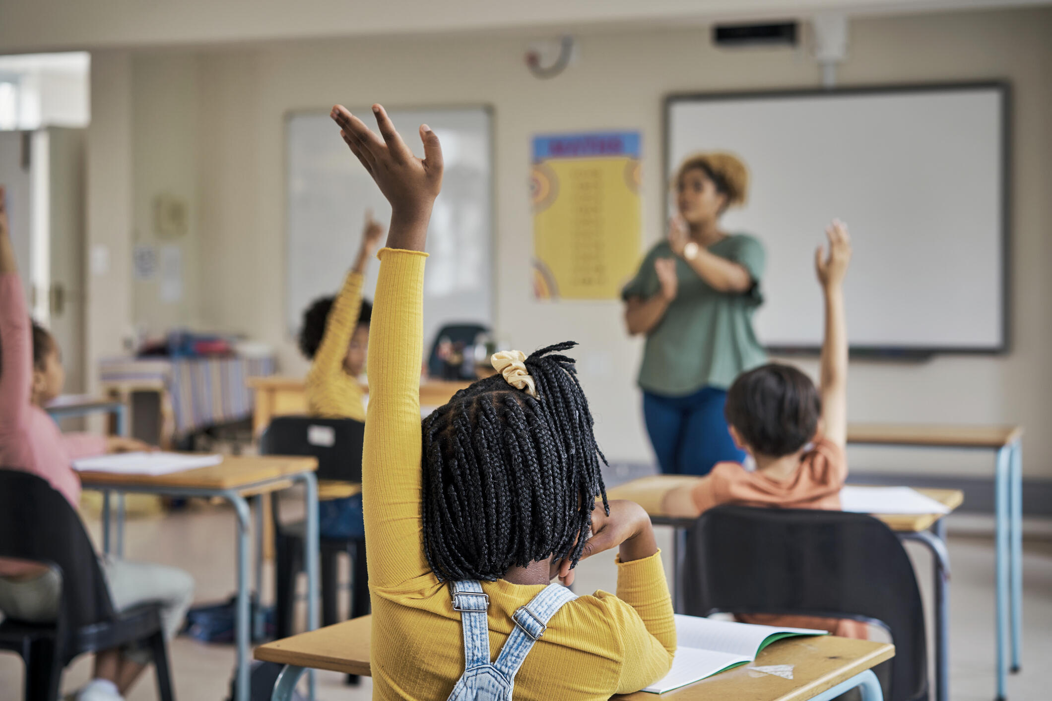 A photo of a classroom with a teacher at the front of the room and several students sitting at desks. The students are all raising their hands. 