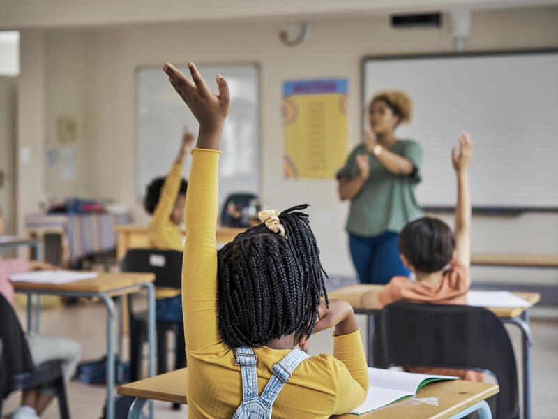 A photo of a classroom with a teacher at the front of the room and several students sitting at desks. The students are all raising their hands. 