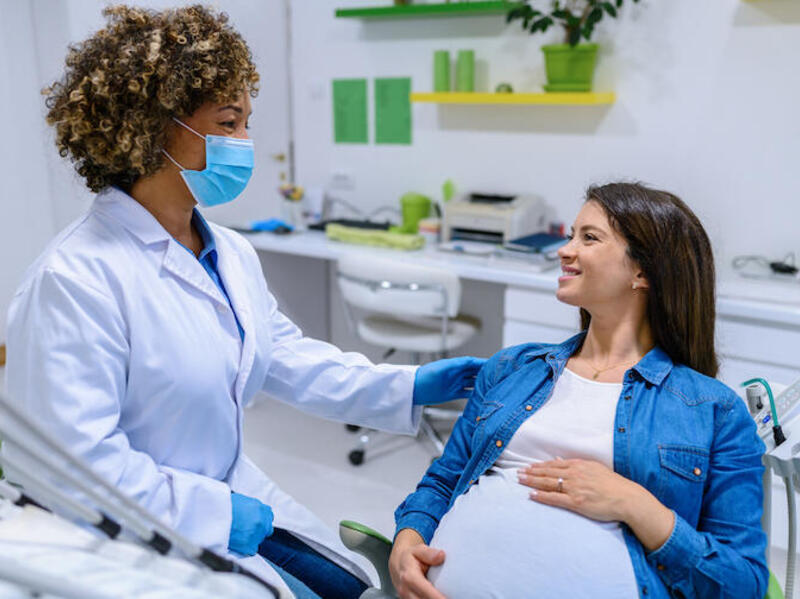 A dentist wearing a face mask, white lab coat, and latex gloves, next to a pregnant women in an exam chair. 