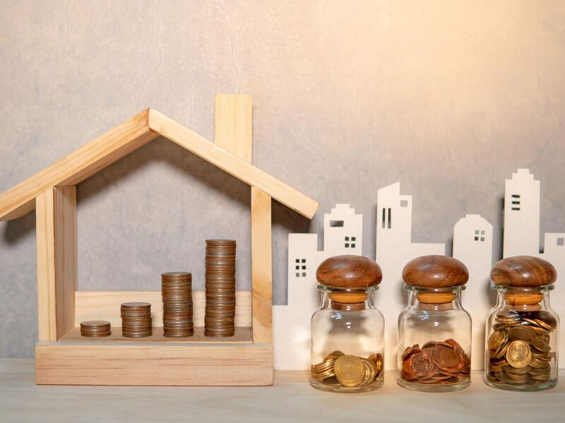 A small wooden house with stacks of coins inside. Next to it are three glass shakers with coins in them. Behind all that is the outlines of a city skyline.
