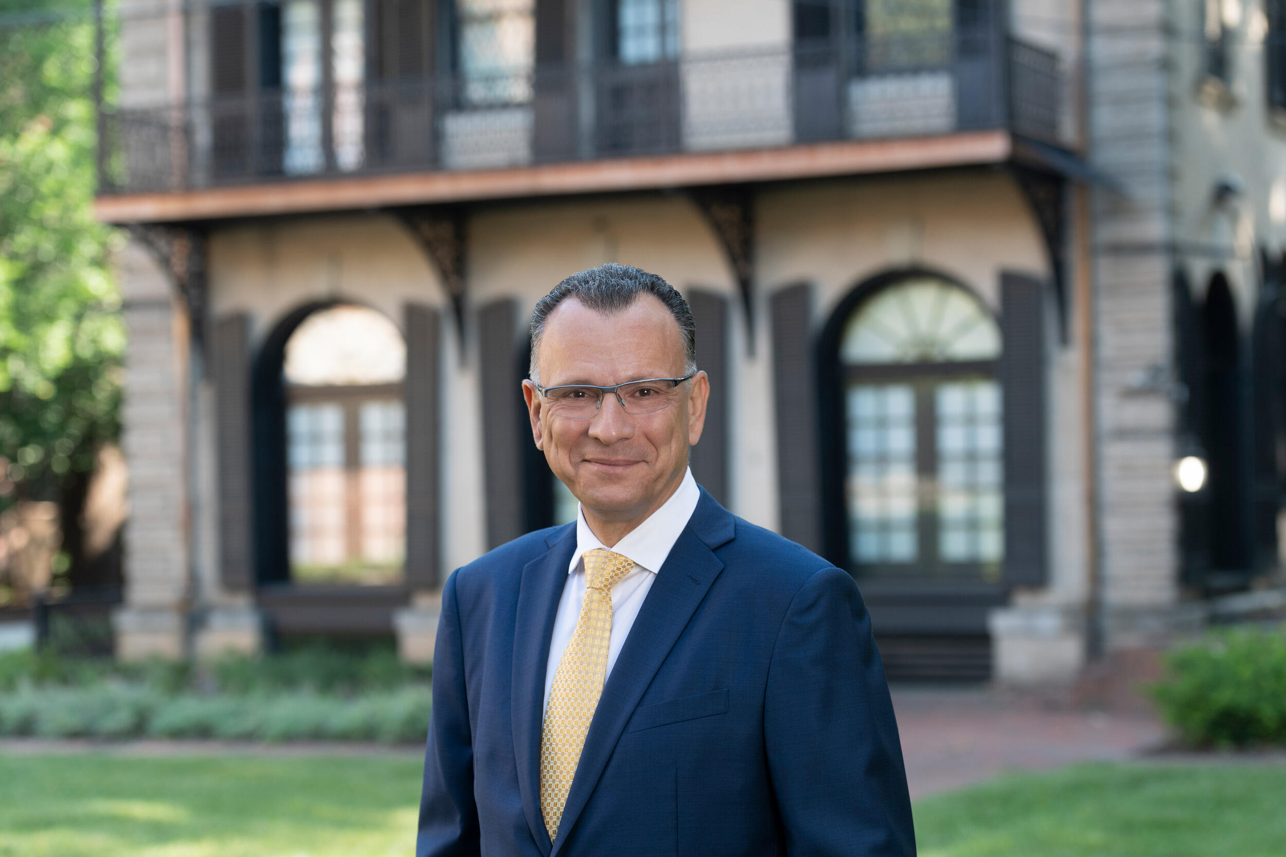 Photo of smiling man in a coat and tie and glasses with a building in the background.