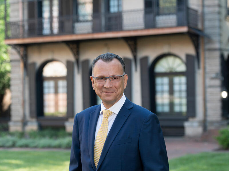 Photo of smiling man in a coat and tie and glasses with a building in the background.
