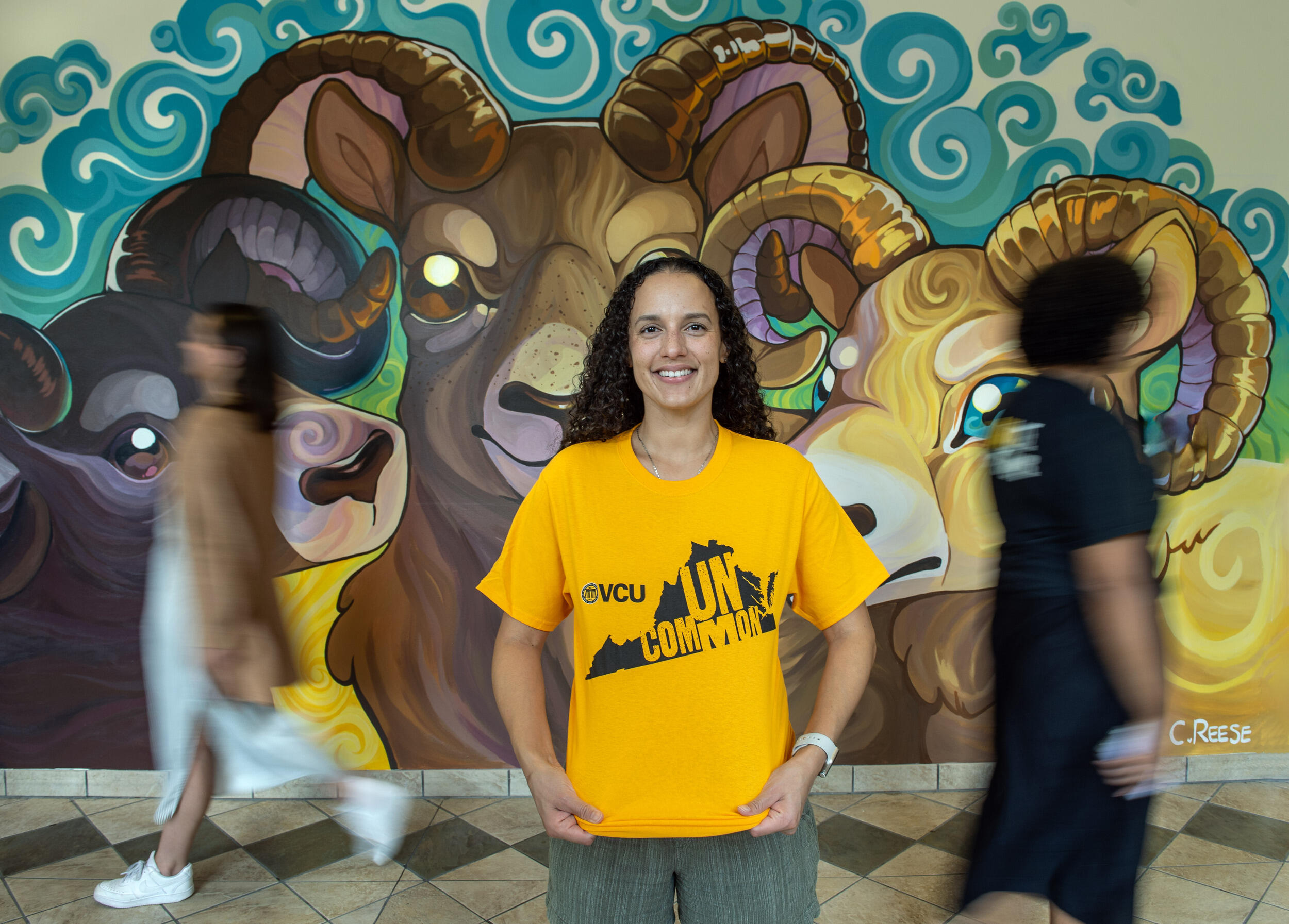 A photo of a woman from the waist up. She is wearing a yellow t-shirt with the shape of Virginia printed on it. Inside the state's silhouette is yellow text that reads \"UNCOMMON.\" Behind the woman are people walking past her and a mural of three rams. 