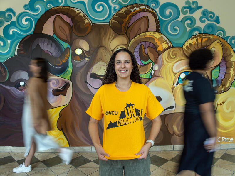 A photo of a woman from the waist up. She is wearing a yellow t-shirt with the shape of Virginia printed on it. Inside the state's silhouette is yellow text that reads \"UNCOMMON.\" Behind the woman are people walking past her and a mural of three rams. 