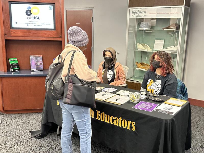 Two people sitting behind a table covered in flyers wearing facemasks. Another person wearing a backpack and back stands in front of the table. 