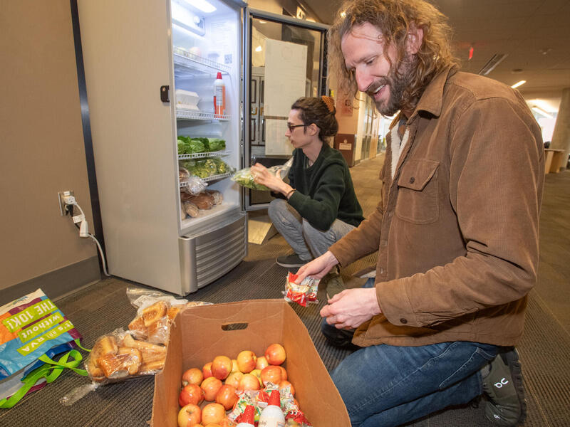 A photo of a man kneeling in front of a box of apples and another man kneeling in front of an open fridge. The man in front of the fridge is stocking it with produce. 