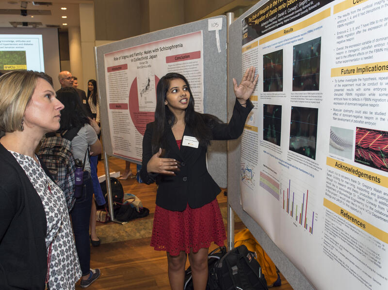 A photo of a two women stanind in front of a poster. The woman on the right is gesturing to the poster while the woman on the left looks at it. 