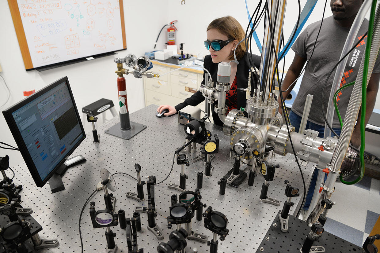 A woman with goggles works on a computer surrounded by lab equipment on a large table. A man in a gray shirt stands to her left.