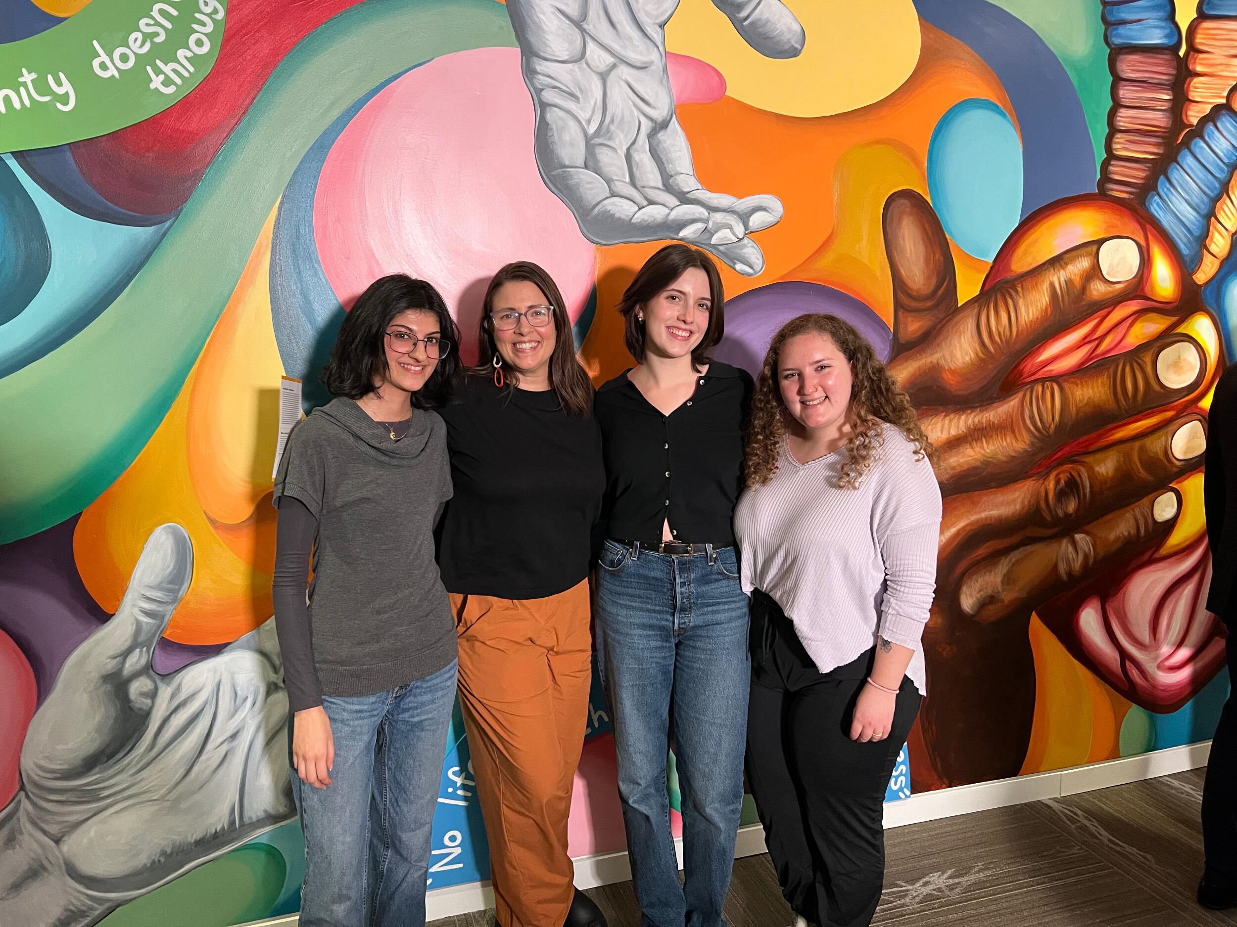 Four women stand together in front of a colorful mural.