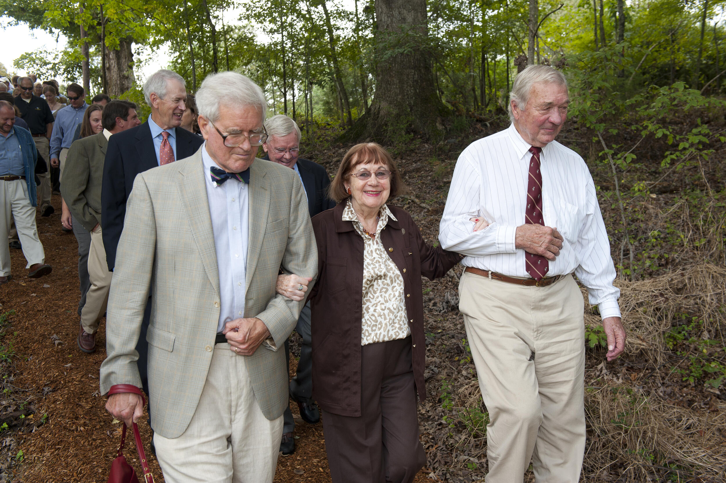 A photo of a crowd of people walking down a path. In the front is a woman who has a man on either side of her holding one of her arms. 