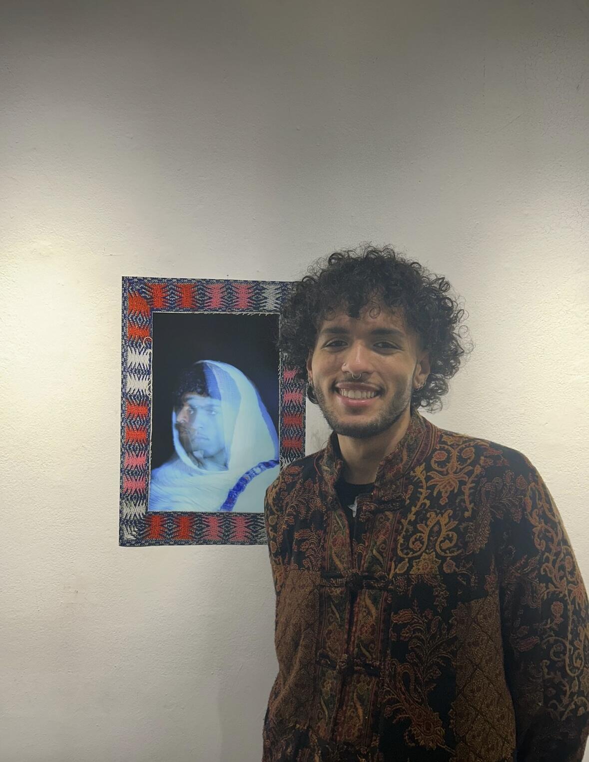 A photo of a man from the waist up. He is standing next to a photograph hanging on a wall