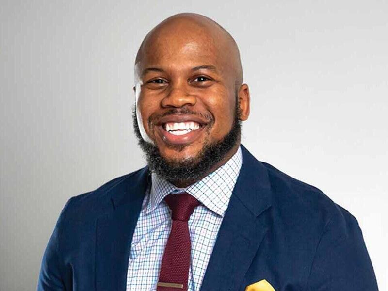 A photo of Deonte Young, sitting and smiling in a suit.