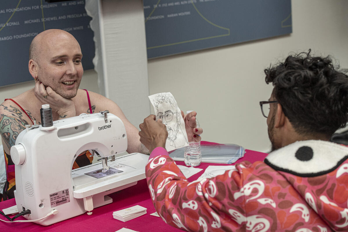 Michael-Birch Pierce sitting behind a sewing machine. A is sitting across from Pierce, holding a five minute portrait made of them. 