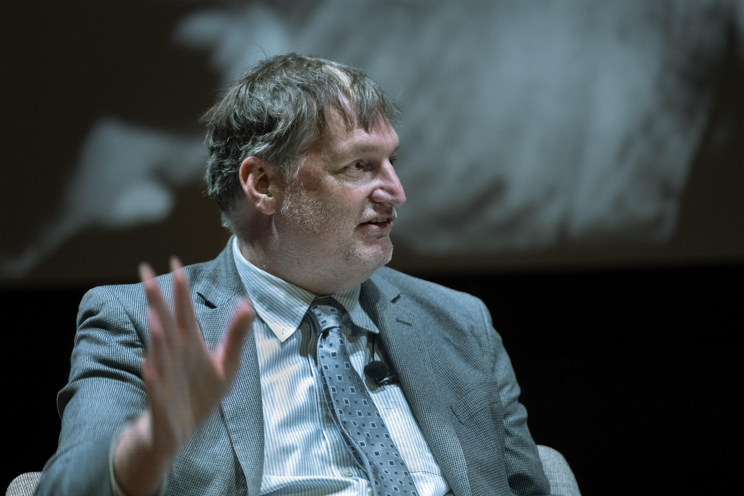 A man wearing a gray sportscoat with his left hand raised looking to the right. 