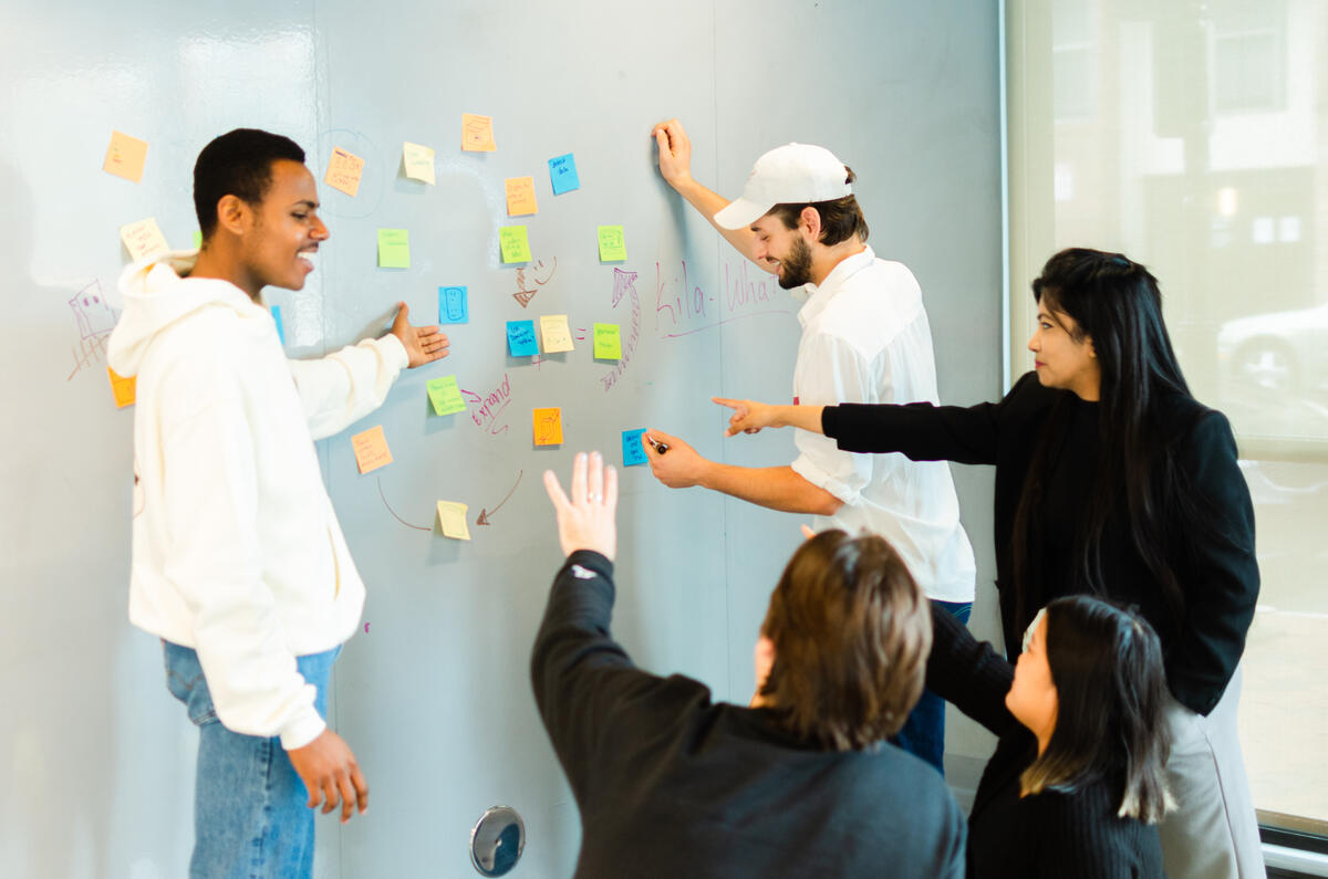 Five people gesturing and looking at a wall with sicky notes on it. 