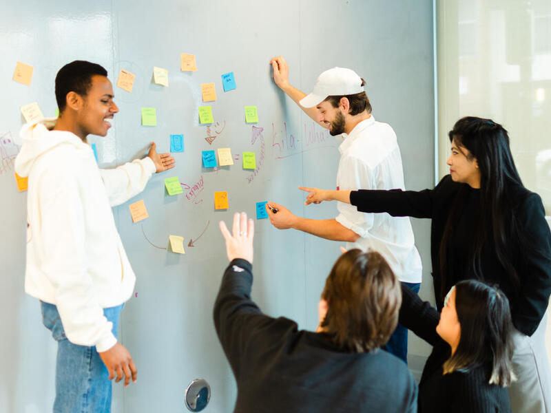 Five people gesturing and looking at a wall with sicky notes on it. 