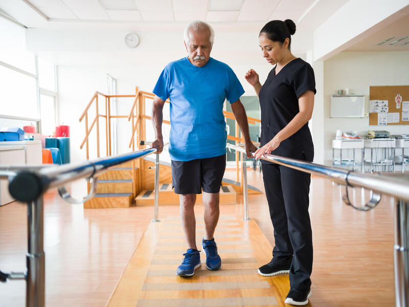 A photo of a man wearing exercise clothing walking between two bars. A woman with black medical scrubs is watching him walk.