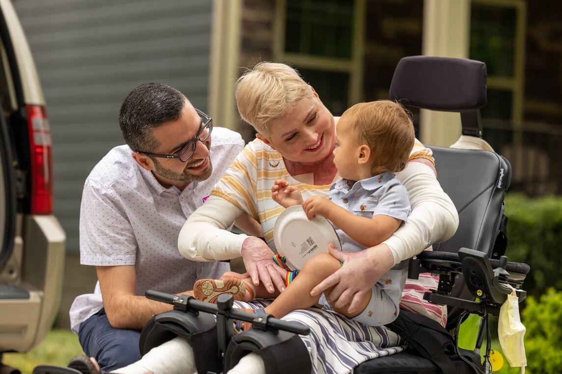 A woman in a wheelchair smiling at a toddler in her lap. A man is leaning next to her also smiling at the child. 
