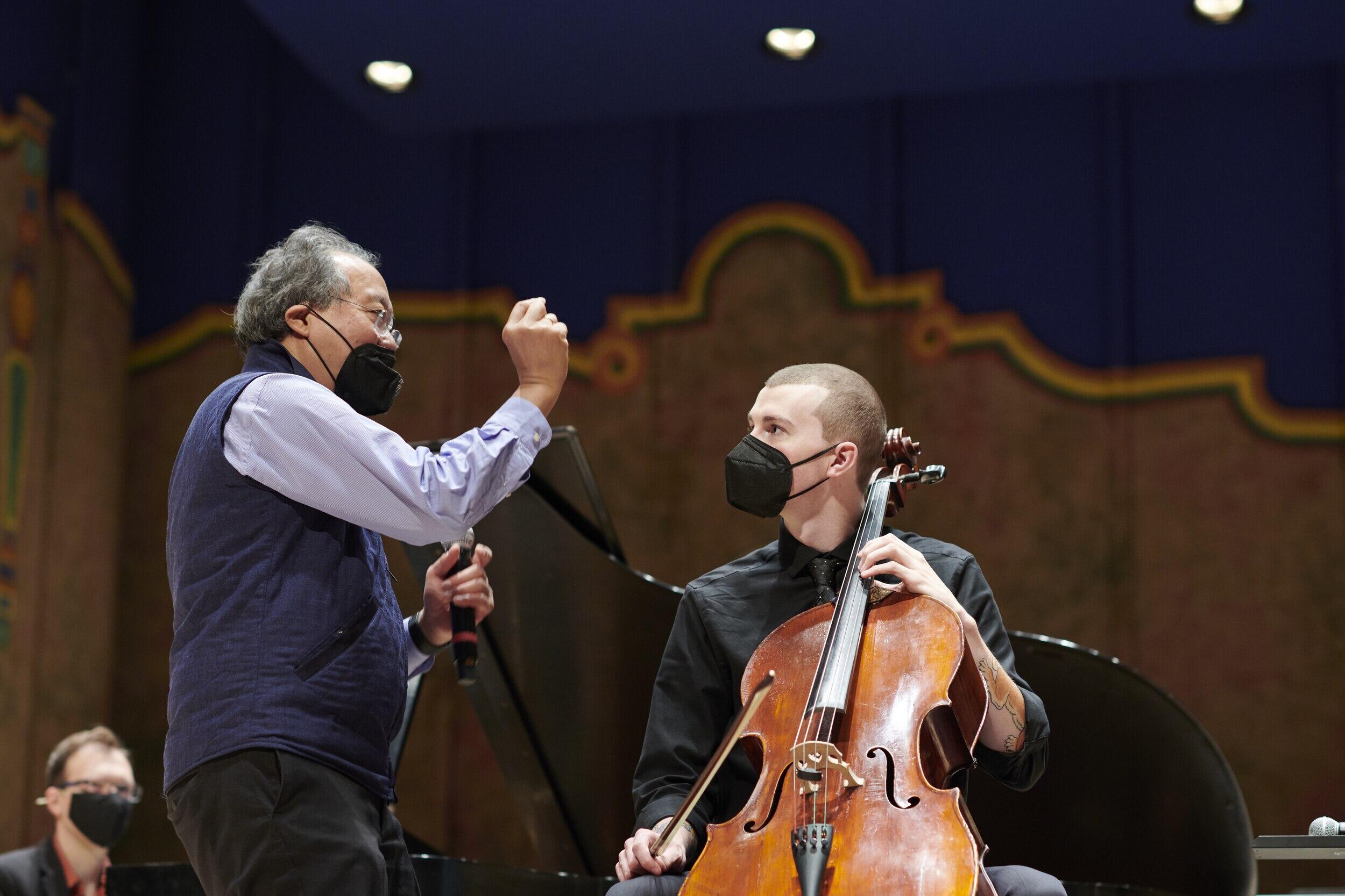 A man wearing a mask standing next to another man who is also wearing a mask and sitting in a chair holding a cello. 
