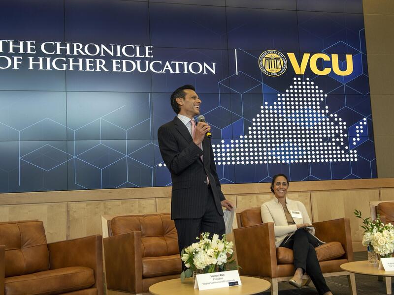 A man standing with a microphone in front of a screen that says \"THE CHRONICLE OF HIGHER EDUCATION\" next to an illustration of Virginia with the letters \"VCU\" above hit. Next to him a woman is sitting in a brown chair and smiling. 