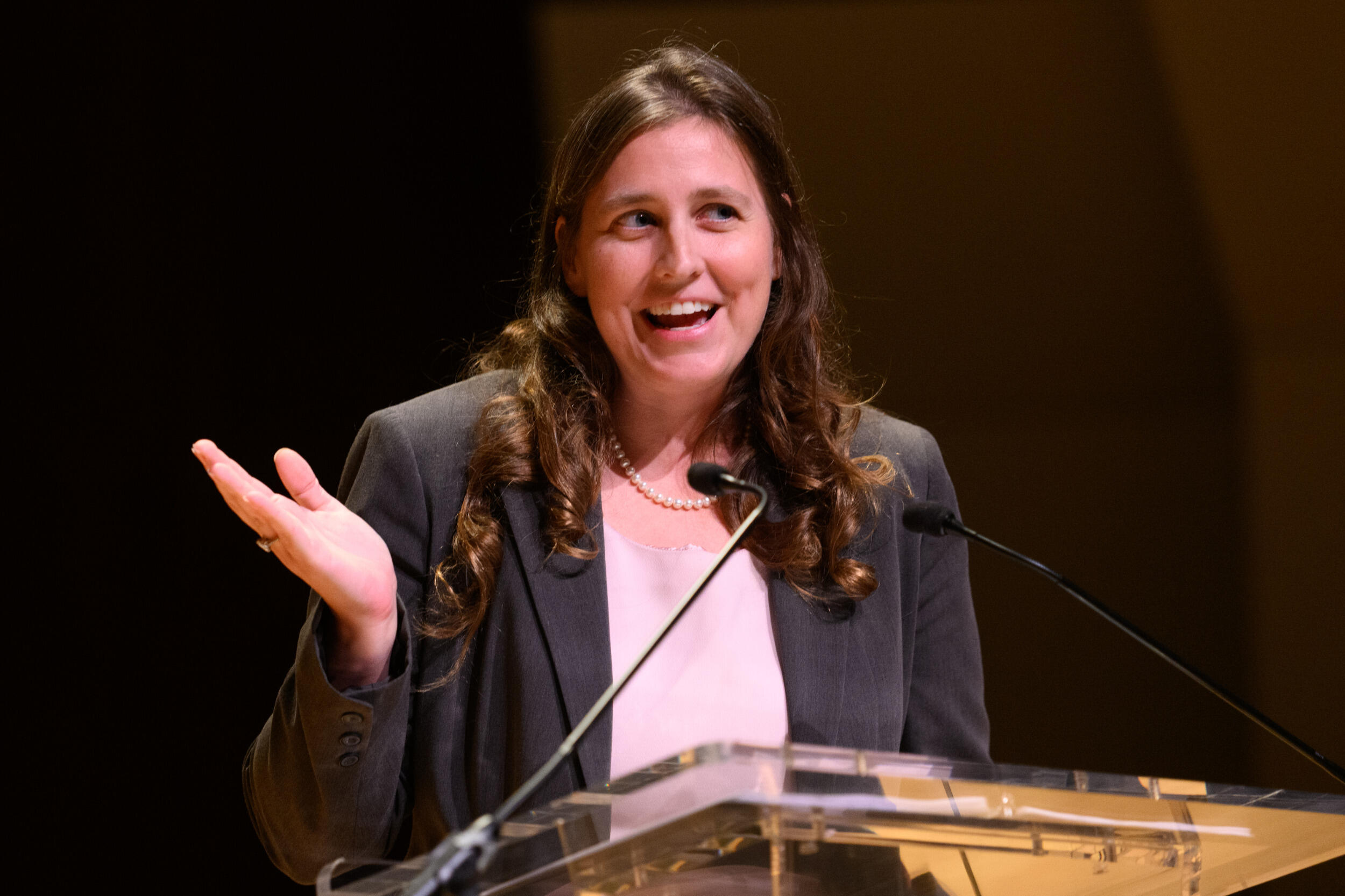 A woman speaks into a microphone at a lectern and gestures with her hand. 