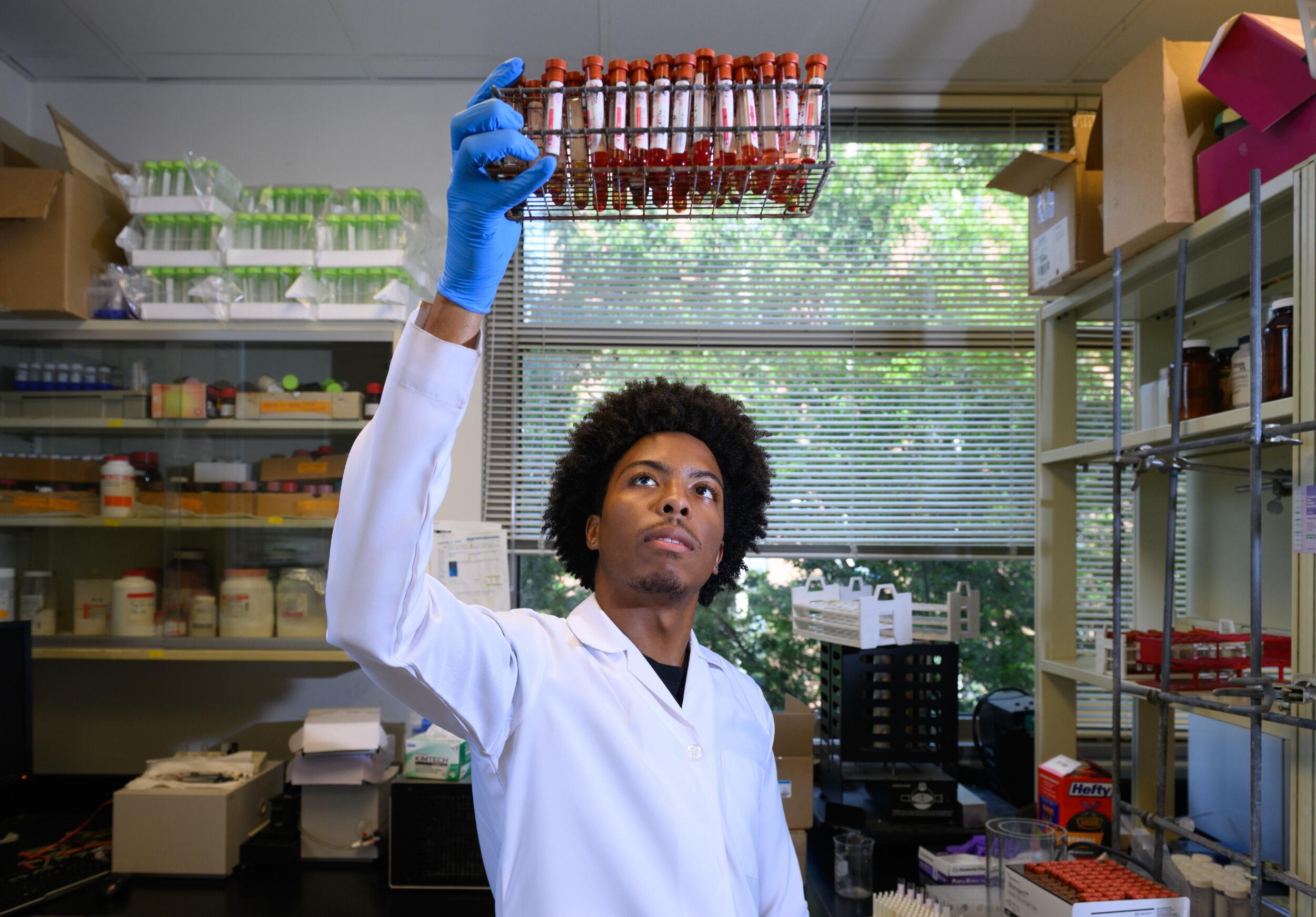 A researcher in a lab coat holds up vials of red liquid.
