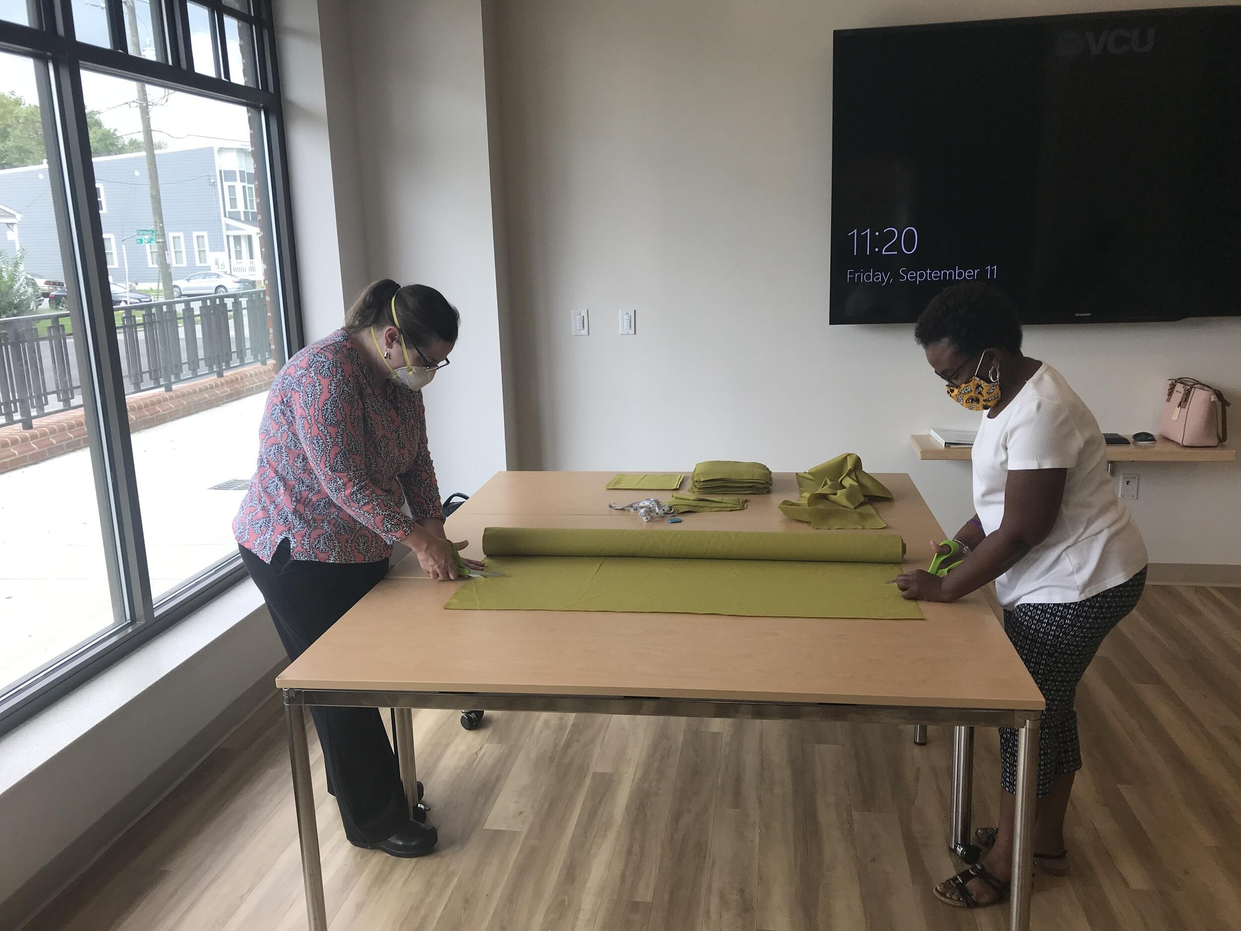 A pair of volunteers cut cloth from a bolt of fabric lying on a table.