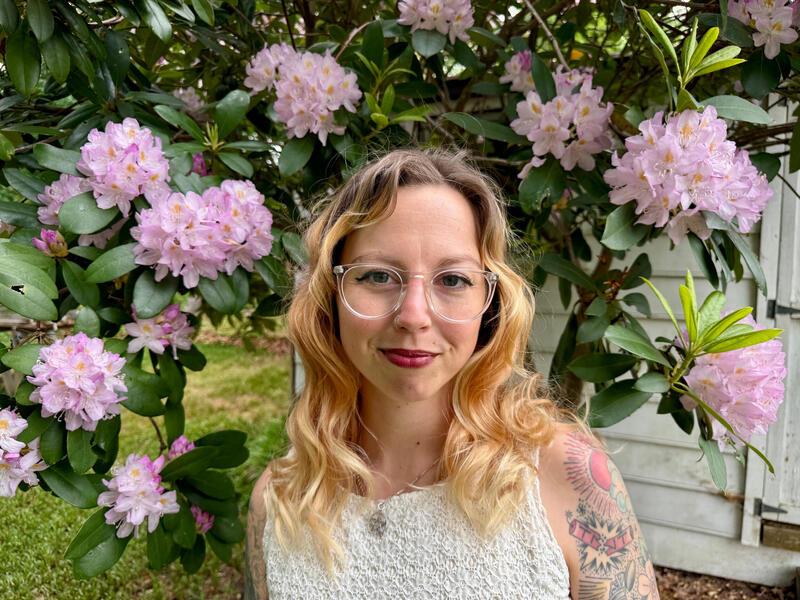 A woman in glasses and a white top stands in front of some flowers.
