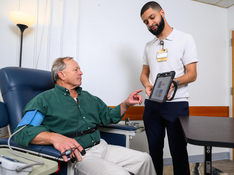 A photo of a man standing and a man sitting. The man standing is showing the sitting man an iPad with information on it. 